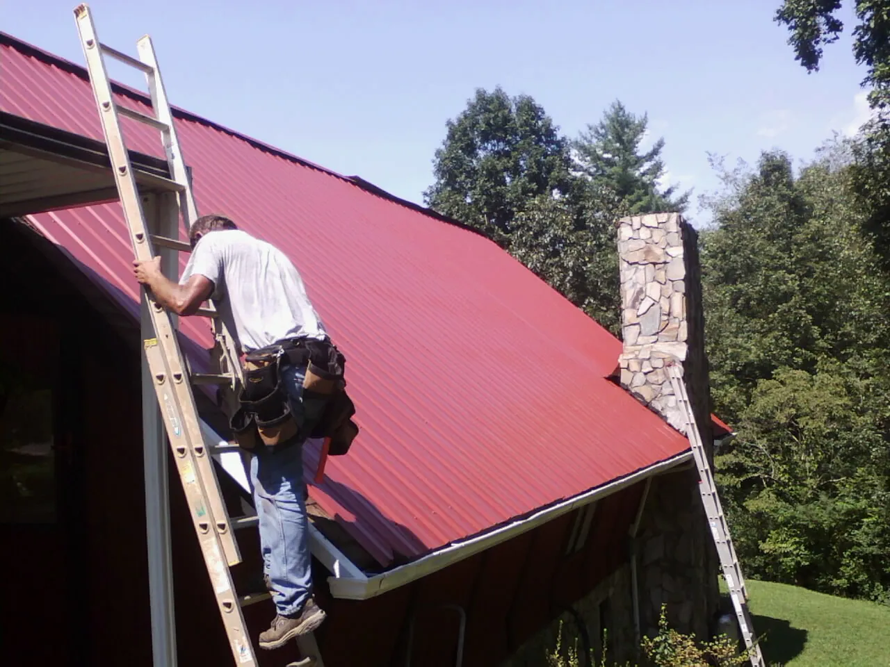 Roofer on a ladder beside a red metal roof and stone chimney — Soffit & Fascia Repair by Roofing Brothers in Crestwood